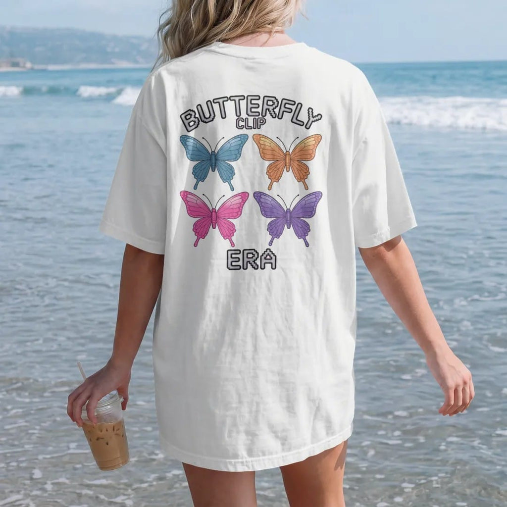 Woman on the beach in a White tee, back view: a row of colorful butterfly hair clips with glitter sparkles, with "BUTTERFLY CLIP" and "ERA" in liquid-chrome bubble letters and small pixel type, printed in glossy gunmetal chrome, cobalt, hot magenta…
