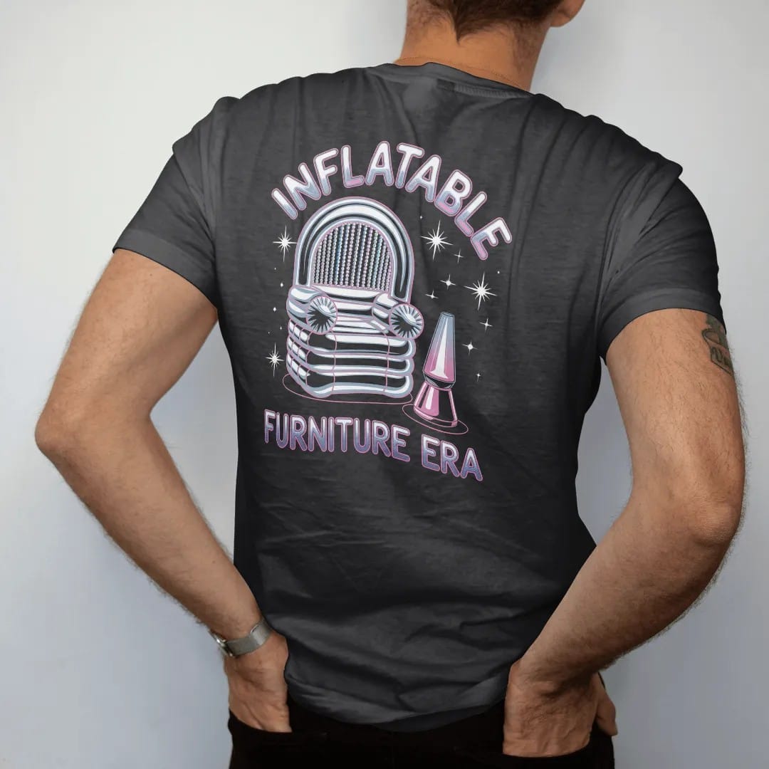 Man wearing a Black tee, seen from the back: a translucent inflatable chair beside a lava lamp and bead curtain, with "INFLATABLE" and "FURNITURE ERA" in liquid-chrome bubble letters and small pixel type, printed in glossy gunmetal chrome, cobalt,…