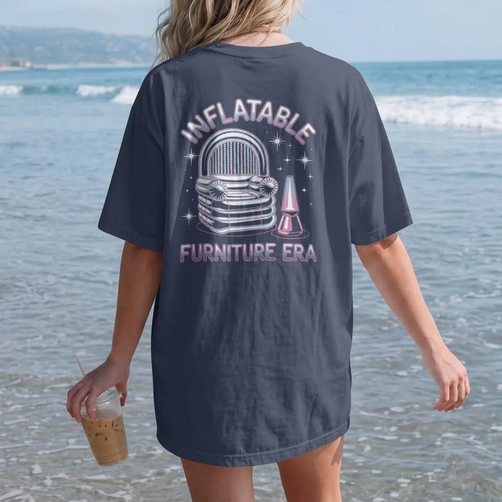 Woman on the beach in a Navy tee, back view: a translucent inflatable chair beside a lava lamp and bead curtain, with "INFLATABLE" and "FURNITURE ERA" in liquid-chrome bubble letters and small pixel type, printed in glossy gunmetal chrome, cobalt,…
