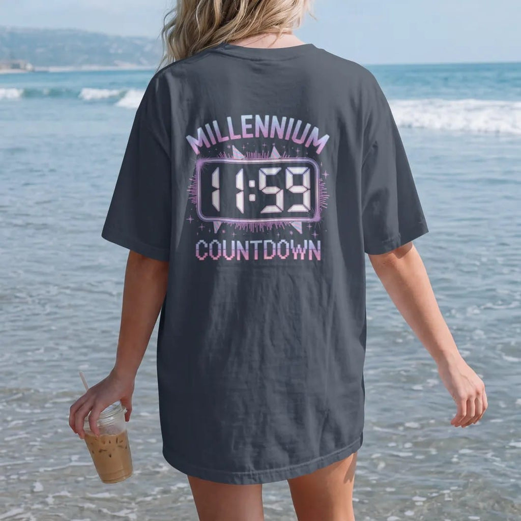 Woman on the beach in a Midnight tee, back view: a glowing digital clock at 11:59 with confetti and party hats, with "MILLENNIUM" and "COUNTDOWN" in liquid-chrome bubble letters and small pixel type, printed in glossy chrome silver, baby blue,…