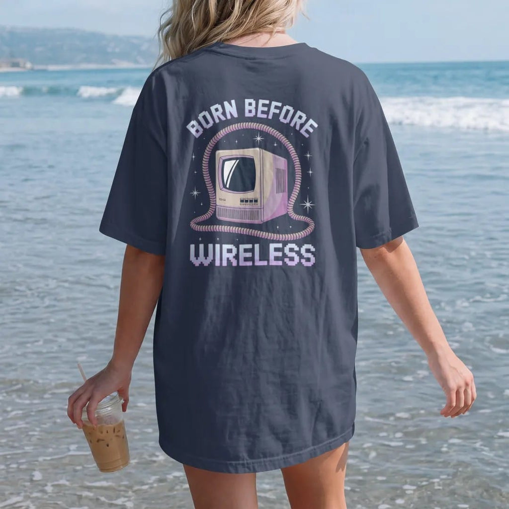 Woman on the beach in a Navy tee, back view: a tangled phone cord framing a beige computer tower, with "BORN BEFORE" and "WIRELESS" in liquid-chrome bubble letters and small pixel type, printed in glossy gunmetal chrome, cobalt, hot magenta and…