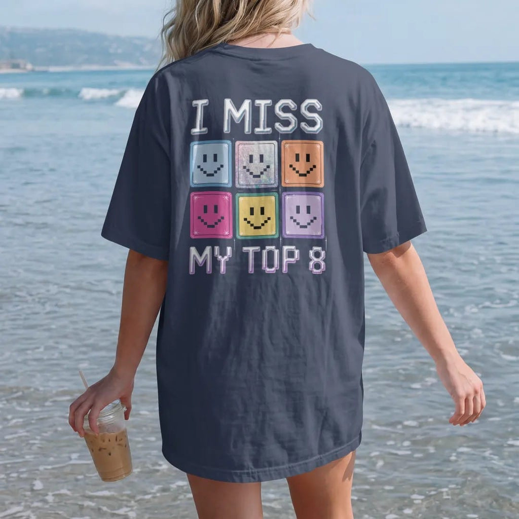 Woman on the beach in a Navy tee, back view: a grid of eight pixel smiley photo frames, with "I MISS" and "MY TOP 8" in liquid-chrome bubble letters and small pixel type, printed in glossy gunmetal chrome, cobalt, hot magenta and violet with…