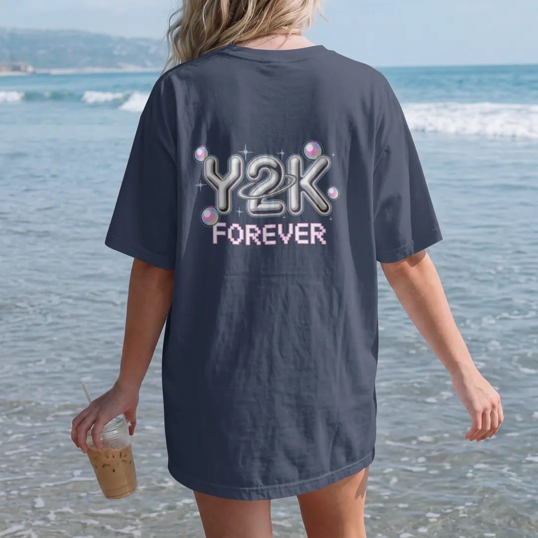 Woman on the beach in a Navy tee, back view: big chrome bubble letters with star sparkles and an orbit ring, with "Y2K" and "FOREVER" in liquid-chrome bubble letters and small pixel type, printed in glossy gunmetal chrome, cobalt, hot magenta and…