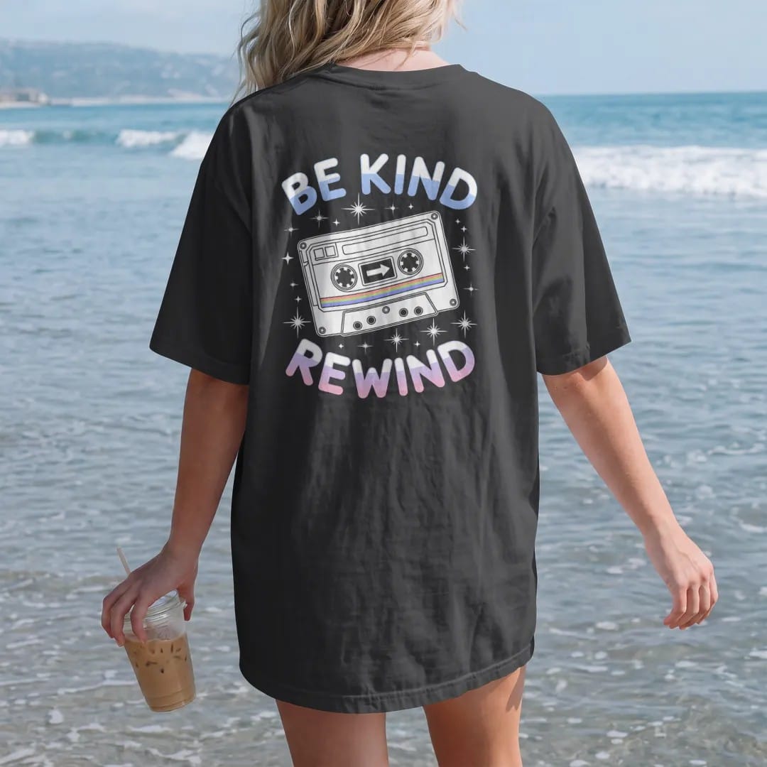 Woman on the beach in a Black tee, back view: a chunky video cassette with a rewind arrow sticker, with "BE KIND" and "REWIND" in liquid-chrome bubble letters and small pixel type, printed in glossy chrome silver, baby blue, bubblegum pink and…