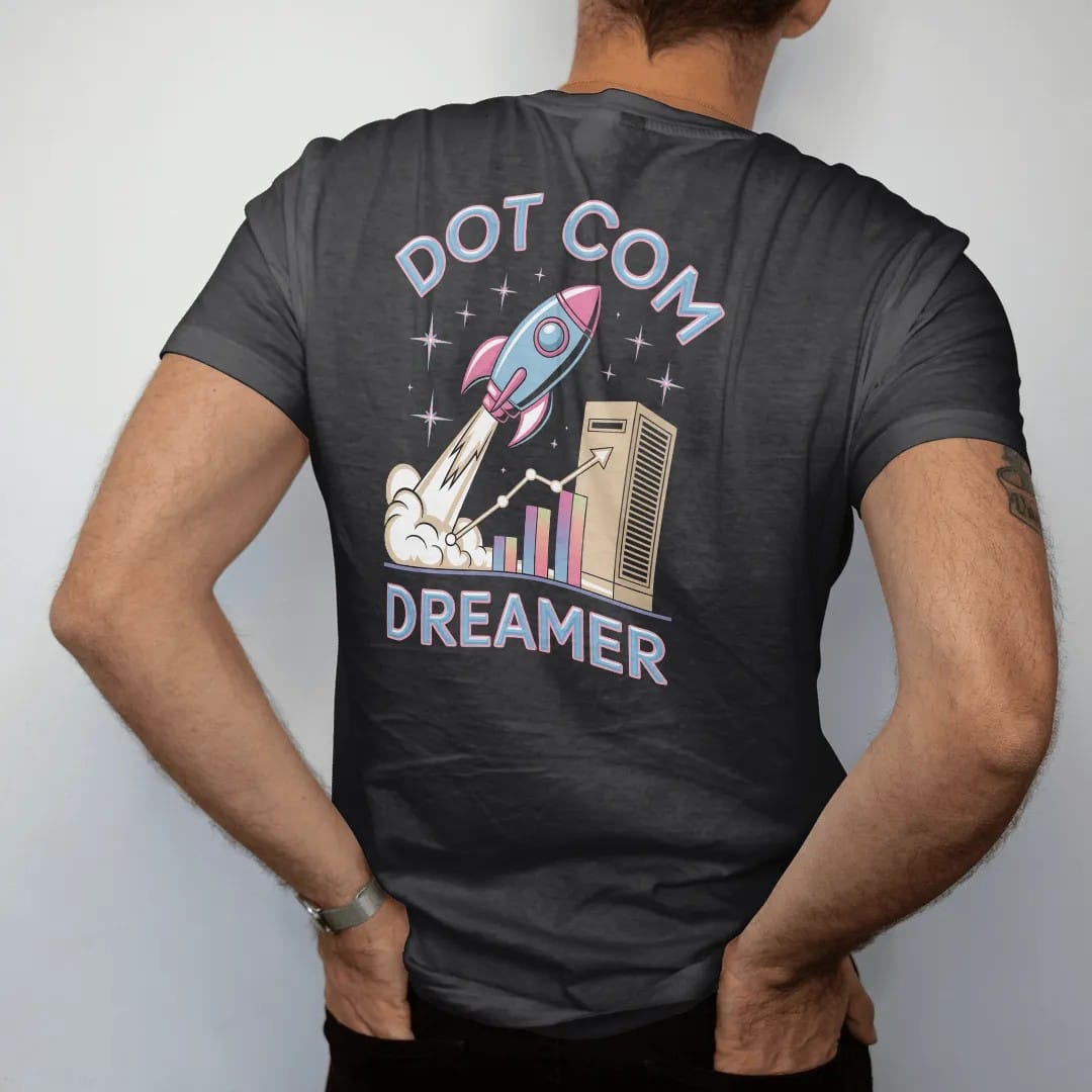 Man wearing a Black tee, seen from the back: a mouse-shaped rocket launching from a beige computer tower, with "DOT COM" and "DREAMER" in liquid-chrome bubble letters and small pixel type, printed in glossy chrome silver, baby blue, bubblegum pink…