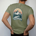 Man wearing a Moss tee, seen from the back: a large arrowhead badge with layered mountains, a pine row and a winding trail, curved header and banner below, faded 70s park-souvenir style, lettered "TRAILHEAD STANDARD / OUTSIDE IS OPEN" in chunky…