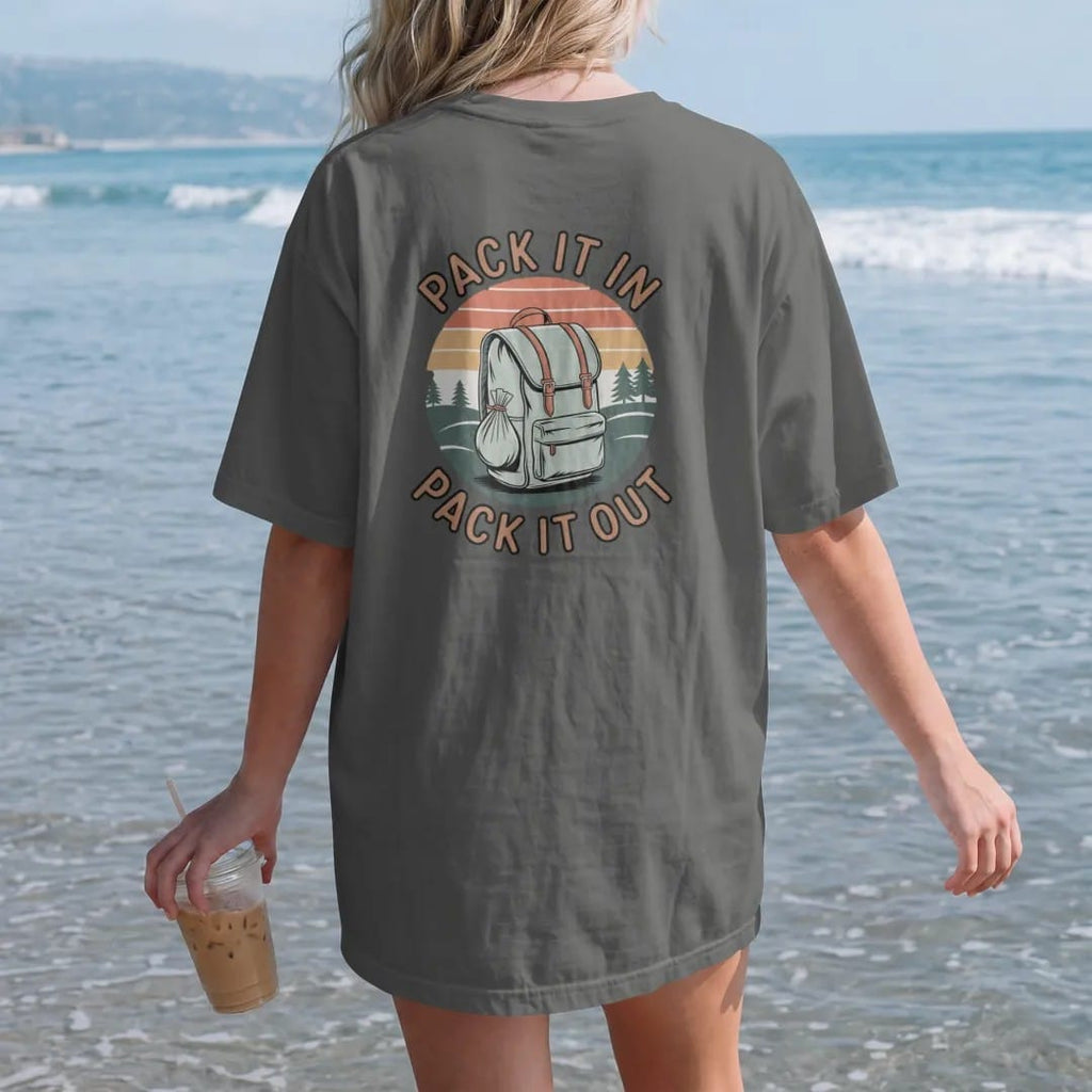 Woman on the beach in a Pepper tee, back view: a loaded backpack with a small trash bag tied on, rolling hills behind, stacked text, faded 70s park-souvenir style, lettered "PACK IT IN / PACK IT OUT" in chunky rounded 70s letters with a drop…
