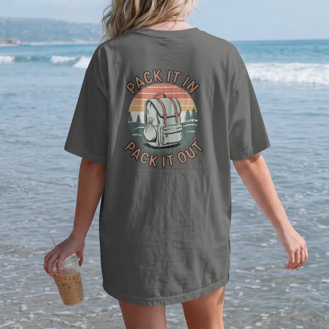 Woman on the beach in a Pepper tee, back view: a loaded backpack with a small trash bag tied on, rolling hills behind, stacked text, faded 70s park-souvenir style, lettered "PACK IT IN / PACK IT OUT" in chunky rounded 70s letters with a drop…