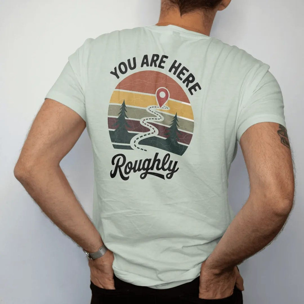 Man wearing a Bay tee, seen from the back: a weathered trail map with a dotted path, little pines and a big red pin, map-board layout, faded 70s park-souvenir style, lettered "YOU ARE HERE / ROUGHLY" in chunky rounded 70s letters with a drop shadow…