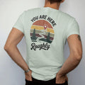 Man wearing a Bay tee, seen from the back: a weathered trail map with a dotted path, little pines and a big red pin, map-board layout, faded 70s park-souvenir style, lettered "YOU ARE HERE / ROUGHLY" in chunky rounded 70s letters with a drop shadow…