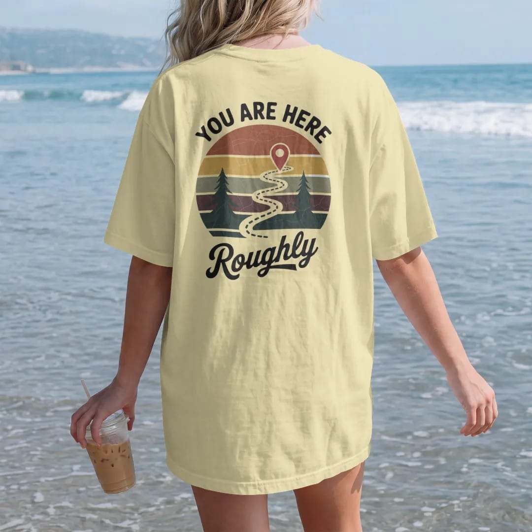 Woman on the beach in a Butter tee, back view: a weathered trail map with a dotted path, little pines and a big red pin, map-board layout, faded 70s park-souvenir style, lettered "YOU ARE HERE / ROUGHLY" in chunky rounded 70s letters with a drop…