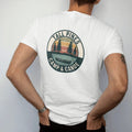 Man wearing a White tee, seen from the back: a round patch with three tall pines over a still lake and a canoe at the shoreline, curved text, faded 70s park-souvenir style, lettered "TALL PINES / CAMP & CANOE" in chunky rounded 70s serif letters…