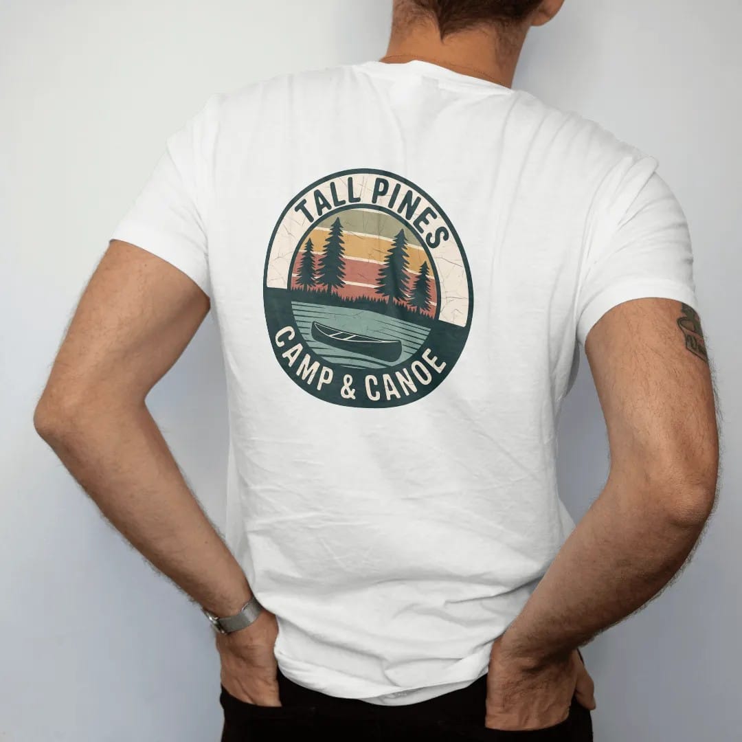 Man wearing a White tee, seen from the back: a round patch with three tall pines over a still lake and a canoe at the shoreline, curved text, faded 70s park-souvenir style, lettered "TALL PINES / CAMP & CANOE" in chunky rounded 70s serif letters…