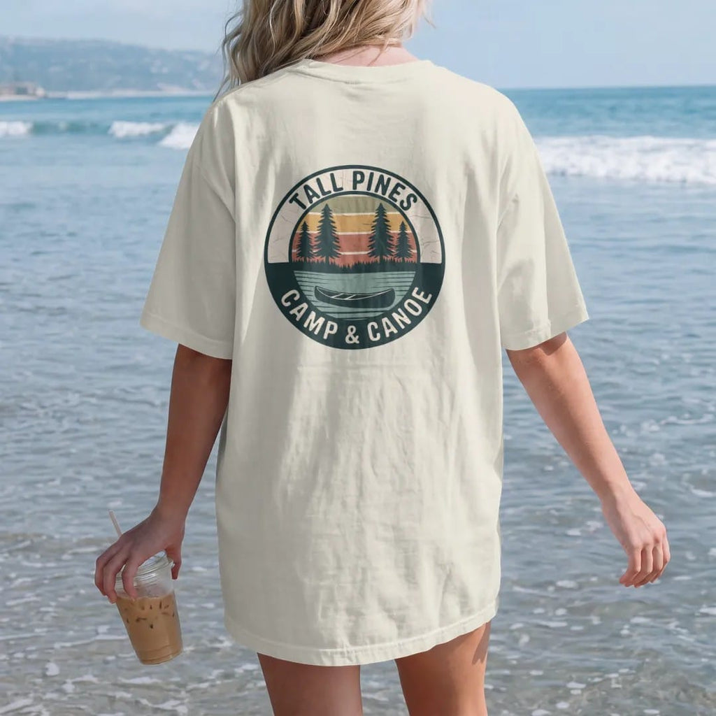 Woman on the beach in a Ivory tee, back view: a round patch with three tall pines over a still lake and a canoe at the shoreline, curved text, faded 70s park-souvenir style, lettered "TALL PINES / CAMP & CANOE" in chunky rounded 70s serif letters…