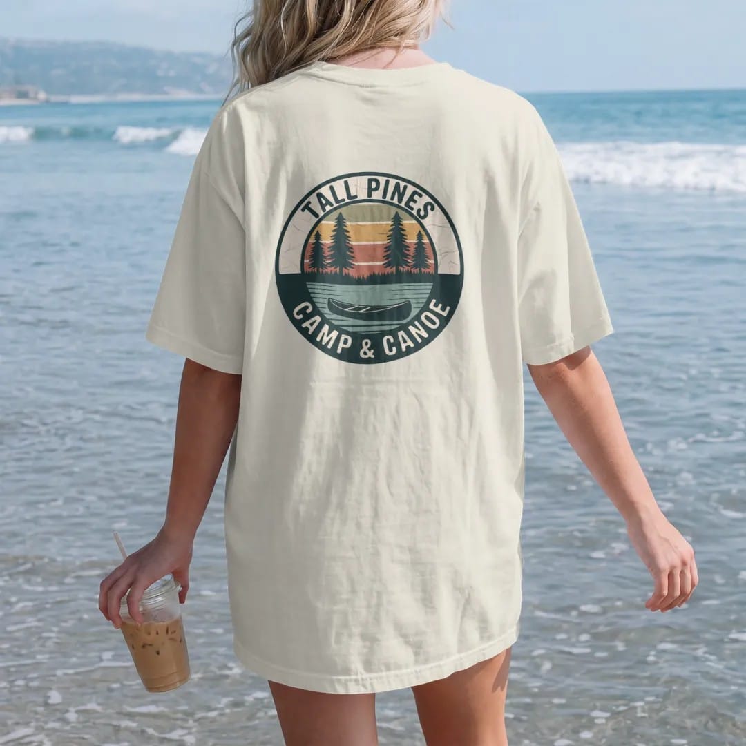 Woman on the beach in a Ivory tee, back view: a round patch with three tall pines over a still lake and a canoe at the shoreline, curved text, faded 70s park-souvenir style, lettered "TALL PINES / CAMP & CANOE" in chunky rounded 70s serif letters…