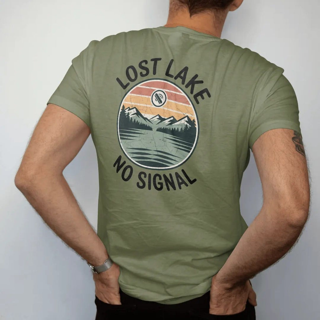 Man wearing a Moss tee, seen from the back: a remote mountain lake in a round frame with a tiny crossed-out phone signal icon in the sky, faded 70s park-souvenir style, lettered "LOST LAKE / NO SIGNAL" in chunky rounded 70s serif letters with a…