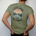 Man wearing a Moss tee, seen from the back: a remote mountain lake in a round frame with a tiny crossed-out phone signal icon in the sky, faded 70s park-souvenir style, lettered "LOST LAKE / NO SIGNAL" in chunky rounded 70s serif letters with a…