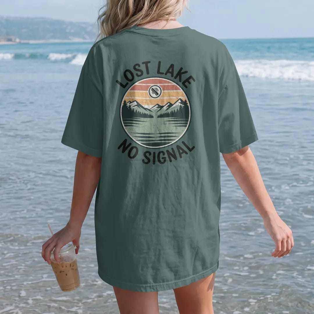 Woman on the beach in a Blue Spruce tee, back view: a remote mountain lake in a round frame with a tiny crossed-out phone signal icon in the sky, faded 70s park-souvenir style, lettered "LOST LAKE / NO SIGNAL" in chunky rounded 70s serif letters…