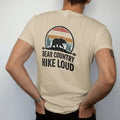 Man wearing a Sandstone tee, seen from the back: a big bear silhouette walking across a ridge with pines, arrowhead frame, warning-sign style, faded 70s park-souvenir style, lettered "BEAR COUNTRY / HIKE LOUD" in chunky rounded 70s letters with a…