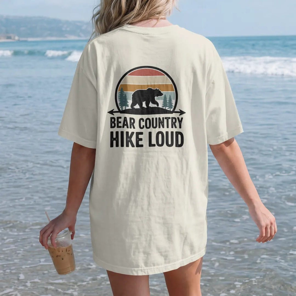 Woman on the beach in a Ivory tee, back view: a big bear silhouette walking across a ridge with pines, arrowhead frame, warning-sign style, faded 70s park-souvenir style, lettered "BEAR COUNTRY / HIKE LOUD" in chunky rounded 70s letters with a drop…