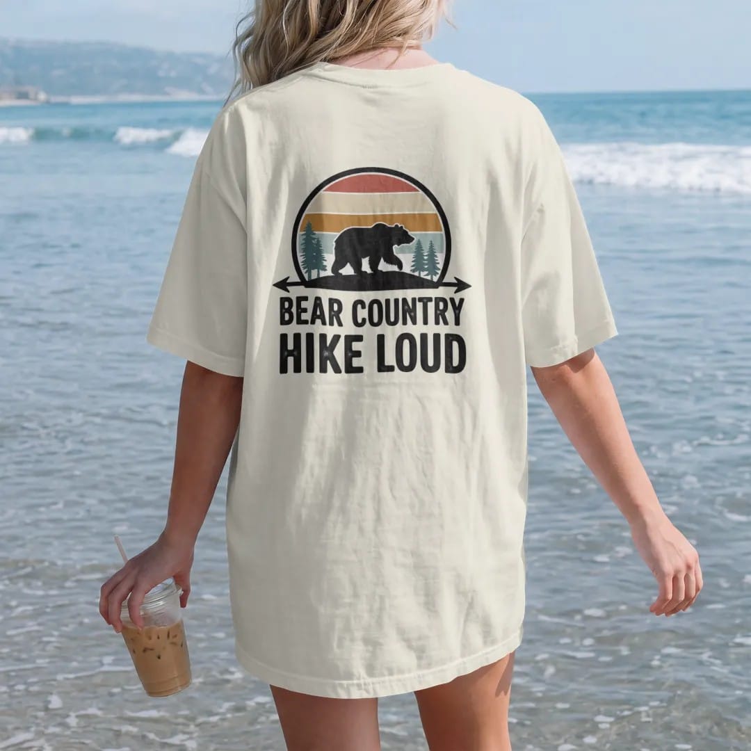 Woman on the beach in a Ivory tee, back view: a big bear silhouette walking across a ridge with pines, arrowhead frame, warning-sign style, faded 70s park-souvenir style, lettered "BEAR COUNTRY / HIKE LOUD" in chunky rounded 70s letters with a drop…