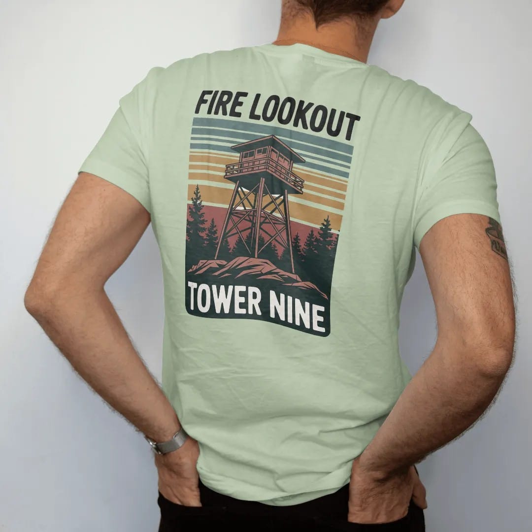 Man wearing a Light Green tee, seen from the back: a tall wooden fire lookout tower on a rocky summit above a sea of pines, vertical poster layout, faded 70s park-souvenir style, lettered "FIRE LOOKOUT / TOWER NINE" in chunky rounded 70s letters…