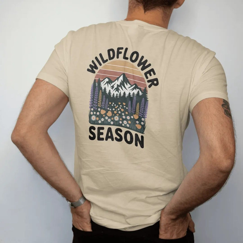Man wearing a Sandstone tee, seen from the back: an alpine meadow bursting with lupine, poppies and daisies below snowy peaks, text arched above, faded 70s park-souvenir style, lettered "WILDFLOWER SEASON" in chunky rounded 70s serif letters with a…