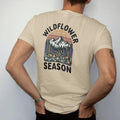 Man wearing a Sandstone tee, seen from the back: an alpine meadow bursting with lupine, poppies and daisies below snowy peaks, text arched above, faded 70s park-souvenir style, lettered "WILDFLOWER SEASON" in chunky rounded 70s serif letters with a…