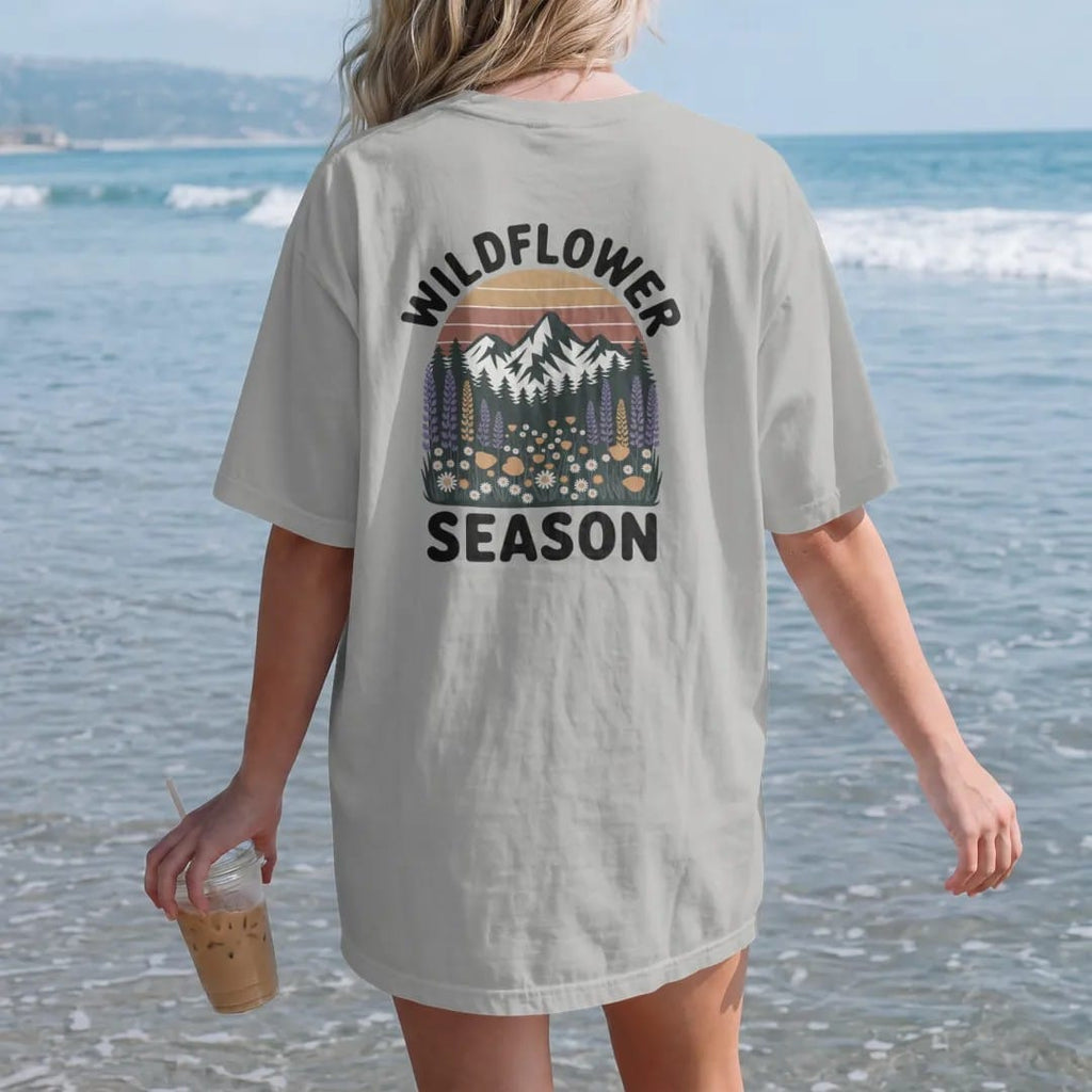 Woman on the beach in a Granite tee, back view: an alpine meadow bursting with lupine, poppies and daisies below snowy peaks, text arched above, faded 70s park-souvenir style, lettered "WILDFLOWER SEASON" in chunky rounded 70s serif letters with a…