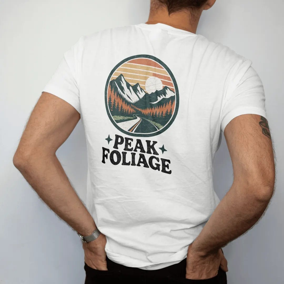 Man wearing a White tee, seen from the back: a mountain valley blazing with orange and gold trees and a winding road, round badge frame, faded 70s park-souvenir style, lettered "PEAK FOLIAGE" in chunky rounded 70s serif letters with a drop shadow,…