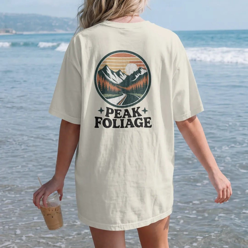 Woman on the beach in a Ivory tee, back view: a mountain valley blazing with orange and gold trees and a winding road, round badge frame, faded 70s park-souvenir style, lettered "PEAK FOLIAGE" in chunky rounded 70s serif letters with a drop shadow,…