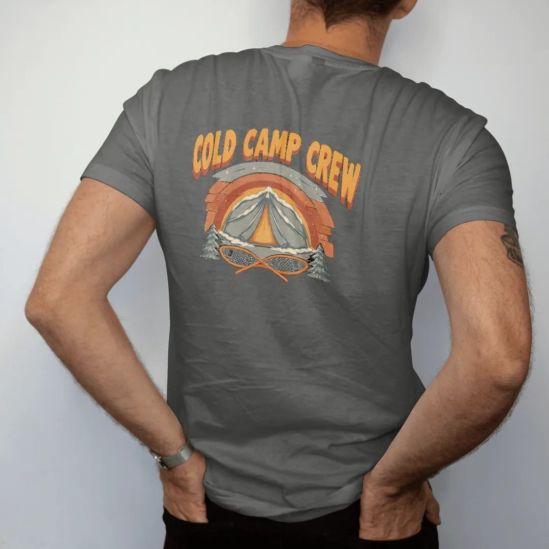 Man wearing a Pepper tee, seen from the back: a snow-covered tent glowing from within under a starry winter sky with crossed snowshoes below, faded 70s park-souvenir style, lettered "COLD CAMP CREW" in chunky rounded 70s letters with a drop shadow,…