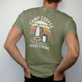 Man wearing a Moss tee, seen from the back: an enamel percolator steaming over a small campfire with a tin mug beside it, label-style layout, faded 70s park-souvenir style, lettered "CAMP COFFEE / BOILED STRONG" in chunky rounded 70s letters with a…