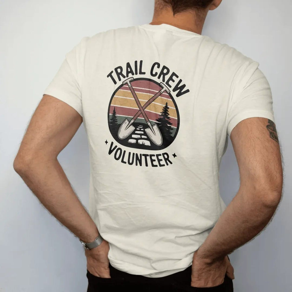 Man wearing a Ivory tee, seen from the back: a crossed shovel and grub hoe over a stone trail step with a small pine, shield badge layout, faded 70s park-souvenir style, lettered "TRAIL CREW / VOLUNTEER" in chunky rounded 70s letters with a drop…