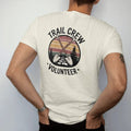 Man wearing a Ivory tee, seen from the back: a crossed shovel and grub hoe over a stone trail step with a small pine, shield badge layout, faded 70s park-souvenir style, lettered "TRAIL CREW / VOLUNTEER" in chunky rounded 70s letters with a drop…