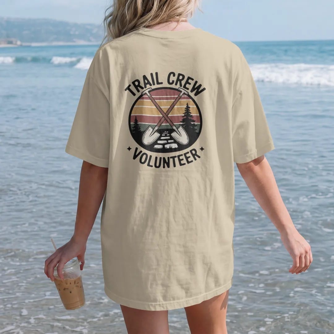 Woman on the beach in a Sandstone tee, back view: a crossed shovel and grub hoe over a stone trail step with a small pine, shield badge layout, faded 70s park-souvenir style, lettered "TRAIL CREW / VOLUNTEER" in chunky rounded 70s letters with a…
