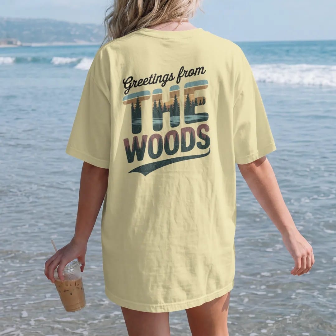 Woman on the beach in a Butter tee, back view: a vintage postcard layout with big letters filled with forest, lake and cabin scenes, faded 70s park-souvenir style, lettered "GREETINGS FROM / THE WOODS" in chunky rounded 70s letters with a drop…