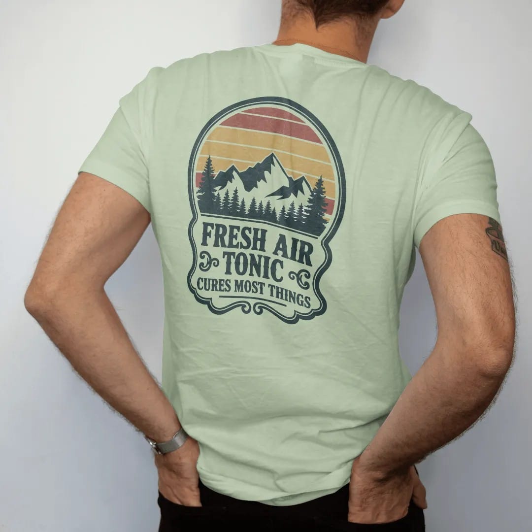 Man wearing a Light Green tee, seen from the back: an old-time medicine bottle label with a mountain scene inside an ornate frame and small scrolls, faded 70s park-souvenir style, lettered "FRESH AIR TONIC / CURES MOST THINGS" in chunky rounded 70s…