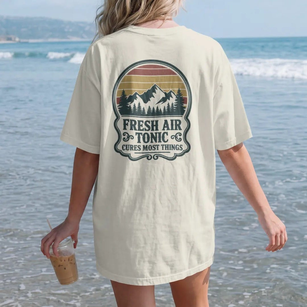 Woman on the beach in a Ivory tee, back view: an old-time medicine bottle label with a mountain scene inside an ornate frame and small scrolls, faded 70s park-souvenir style, lettered "FRESH AIR TONIC / CURES MOST THINGS" in chunky rounded 70s…