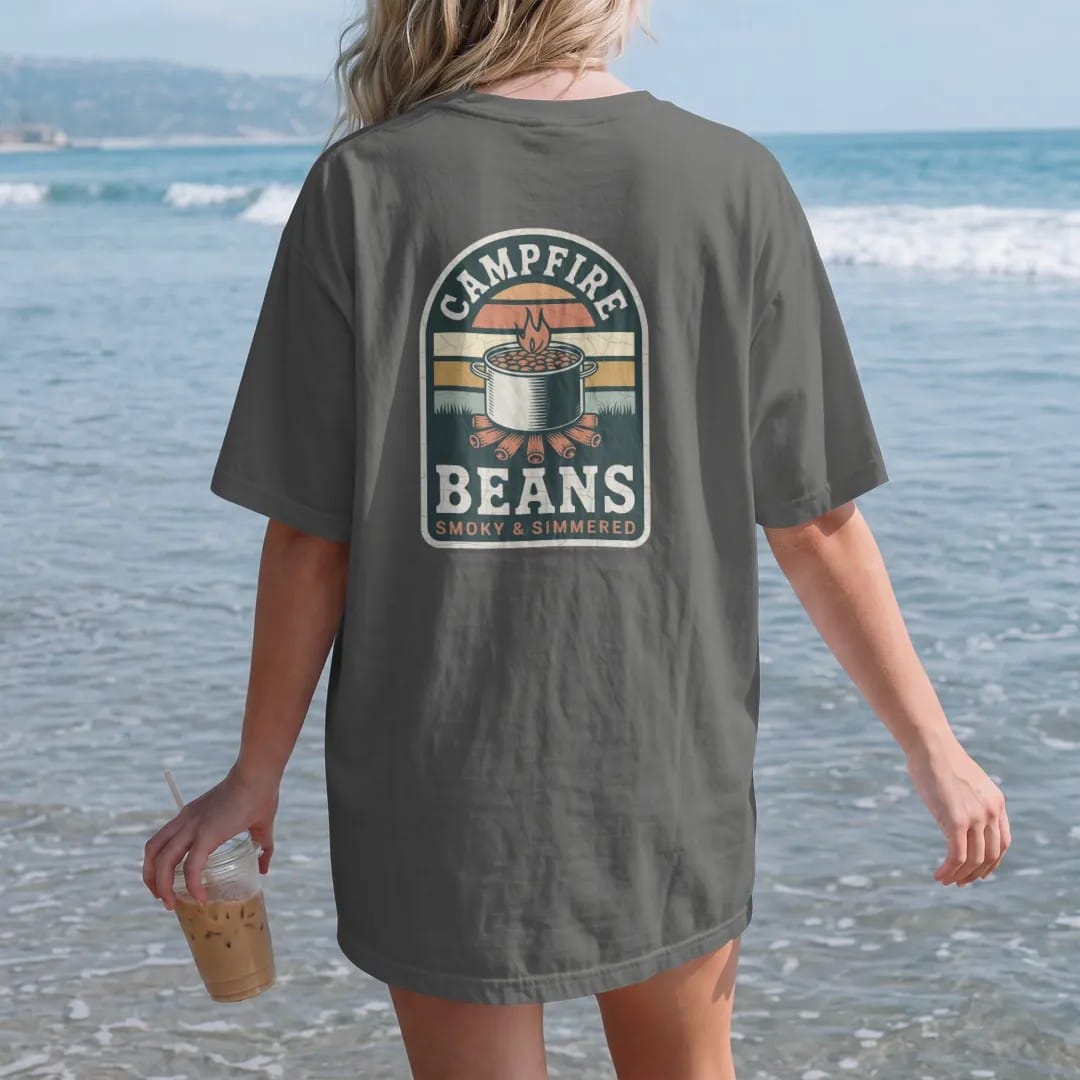 Woman on the beach in a Pepper tee, back view: a retro tin can label with a pot of beans over a fire and a cowboy-style starburst, faded 70s park-souvenir style, lettered "CAMPFIRE BEANS / SMOKY & SIMMERED" in chunky rounded 70s serif letters with…