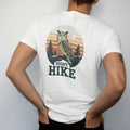 Man wearing a White tee, seen from the back: a great horned owl perched on a pine branch under a full moon with a faint trail below, faded 70s park-souvenir style, lettered "NIGHT HIKE" in chunky rounded 70s letters with a drop shadow, printed in…