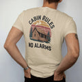 Man wearing a Sandstone tee, seen from the back: a log cabin with smoke curling from the chimney and a wooden sign hanging off the porch, faded 70s park-souvenir style, lettered "CABIN RULES / NO ALARMS" in chunky rounded 70s letters with a drop…