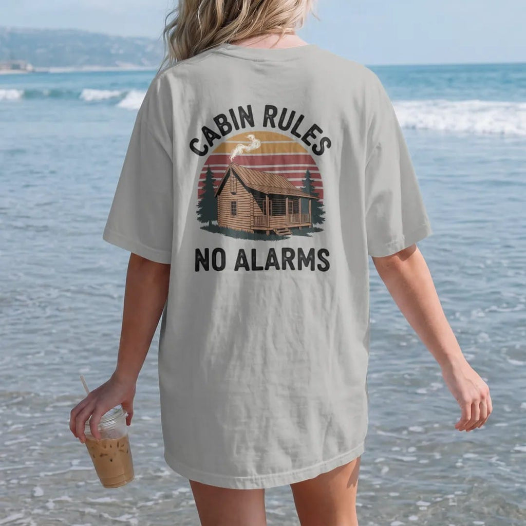 Woman on the beach in a Granite tee, back view: a log cabin with smoke curling from the chimney and a wooden sign hanging off the porch, faded 70s park-souvenir style, lettered "CABIN RULES / NO ALARMS" in chunky rounded 70s letters with a drop…
