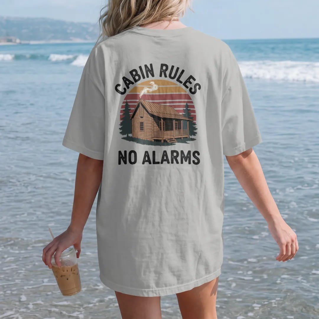 Woman on the beach in a Granite tee, back view: a log cabin with smoke curling from the chimney and a wooden sign hanging off the porch, faded 70s park-souvenir style, lettered "CABIN RULES / NO ALARMS" in chunky rounded 70s letters with a drop…