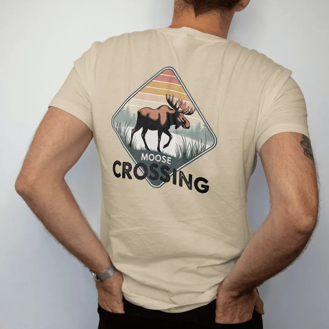 Man wearing a Sandstone tee, seen from the back: a big bull moose stepping across a misty marsh with reeds, diamond road-sign layout, faded 70s park-souvenir style, lettered "MOOSE CROSSING" in chunky rounded 70s letters with a drop shadow, printed…