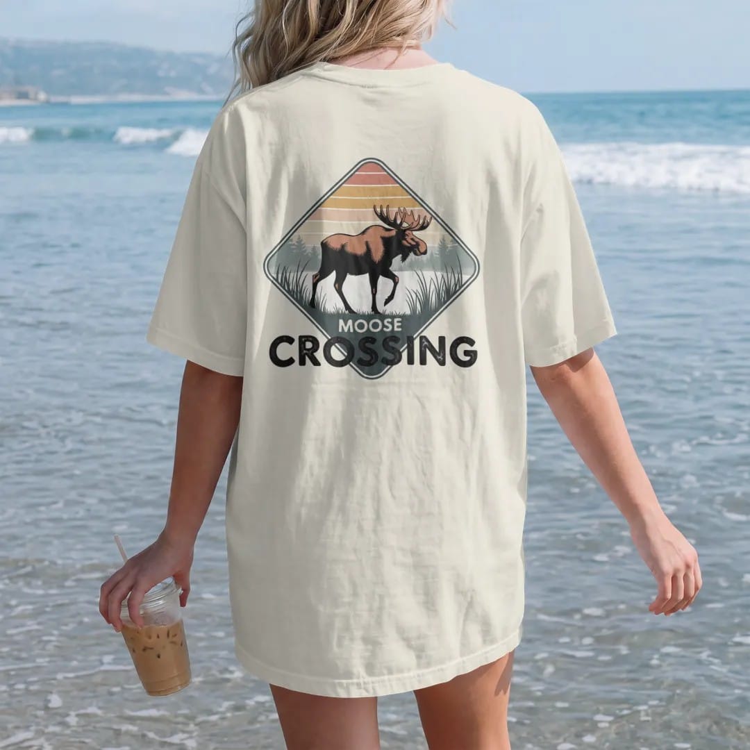 Woman on the beach in a Ivory tee, back view: a big bull moose stepping across a misty marsh with reeds, diamond road-sign layout, faded 70s park-souvenir style, lettered "MOOSE CROSSING" in chunky rounded 70s letters with a drop shadow, printed in…