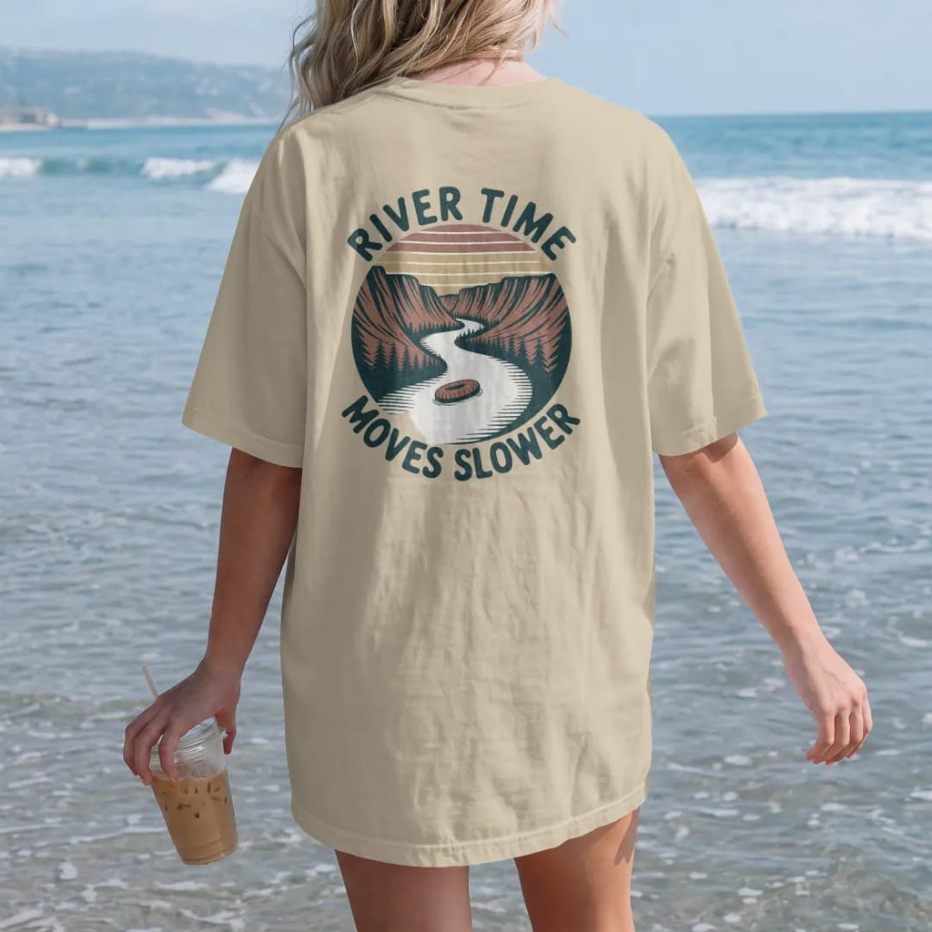 Woman on the beach in a Sandstone tee, back view: a slow winding river through a canyon with a lone tube floater, wavy text following the river, faded 70s park-souvenir style, lettered "RIVER TIME / MOVES SLOWER" in chunky rounded 70s serif letters…
