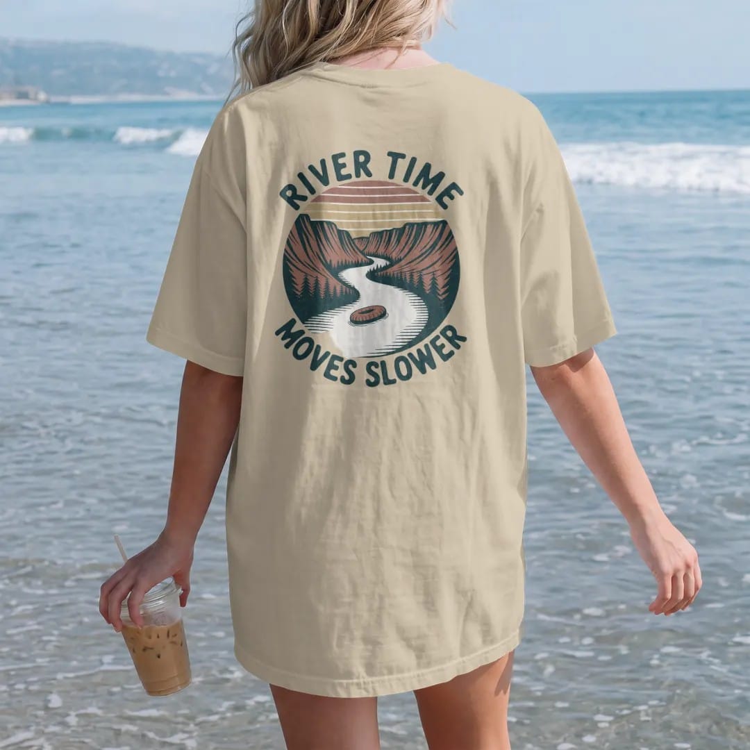 Woman on the beach in a Sandstone tee, back view: a slow winding river through a canyon with a lone tube floater, wavy text following the river, faded 70s park-souvenir style, lettered "RIVER TIME / MOVES SLOWER" in chunky rounded 70s serif letters…