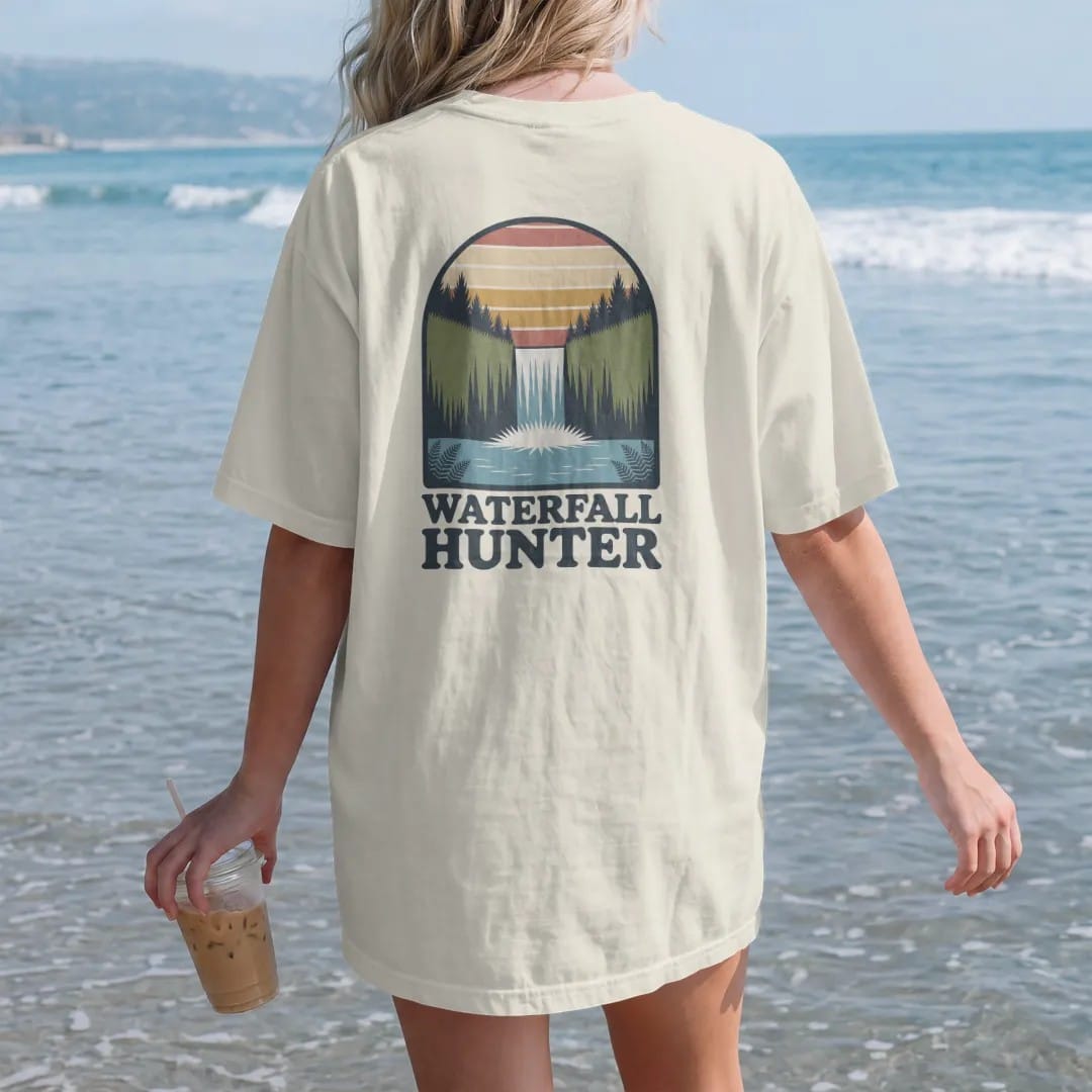 Woman on the beach in a Ivory tee, back view: a tall waterfall pouring into a mossy pool framed by ferns, arched header above, faded 70s park-souvenir style, lettered "WATERFALL HUNTER" in chunky rounded 70s letters with a drop shadow, printed in…