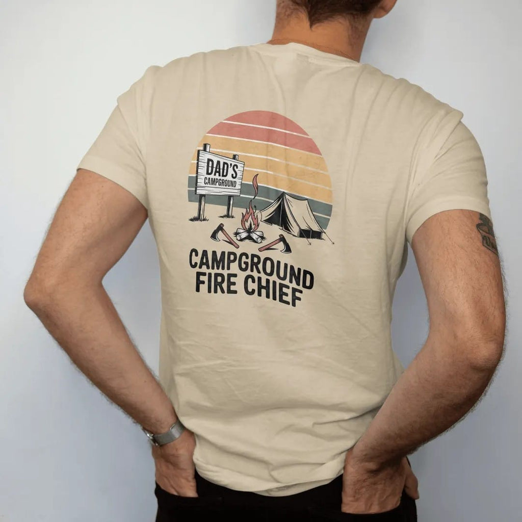 Man wearing a Sandstone tee, seen from the back: a wooden campground entrance sign with a smoking campfire, a tent and crossed hatchets, faded 70s park-souvenir style, lettered "DAD'S CAMPGROUND / FIRE CHIEF" in chunky rounded 70s letters with a…